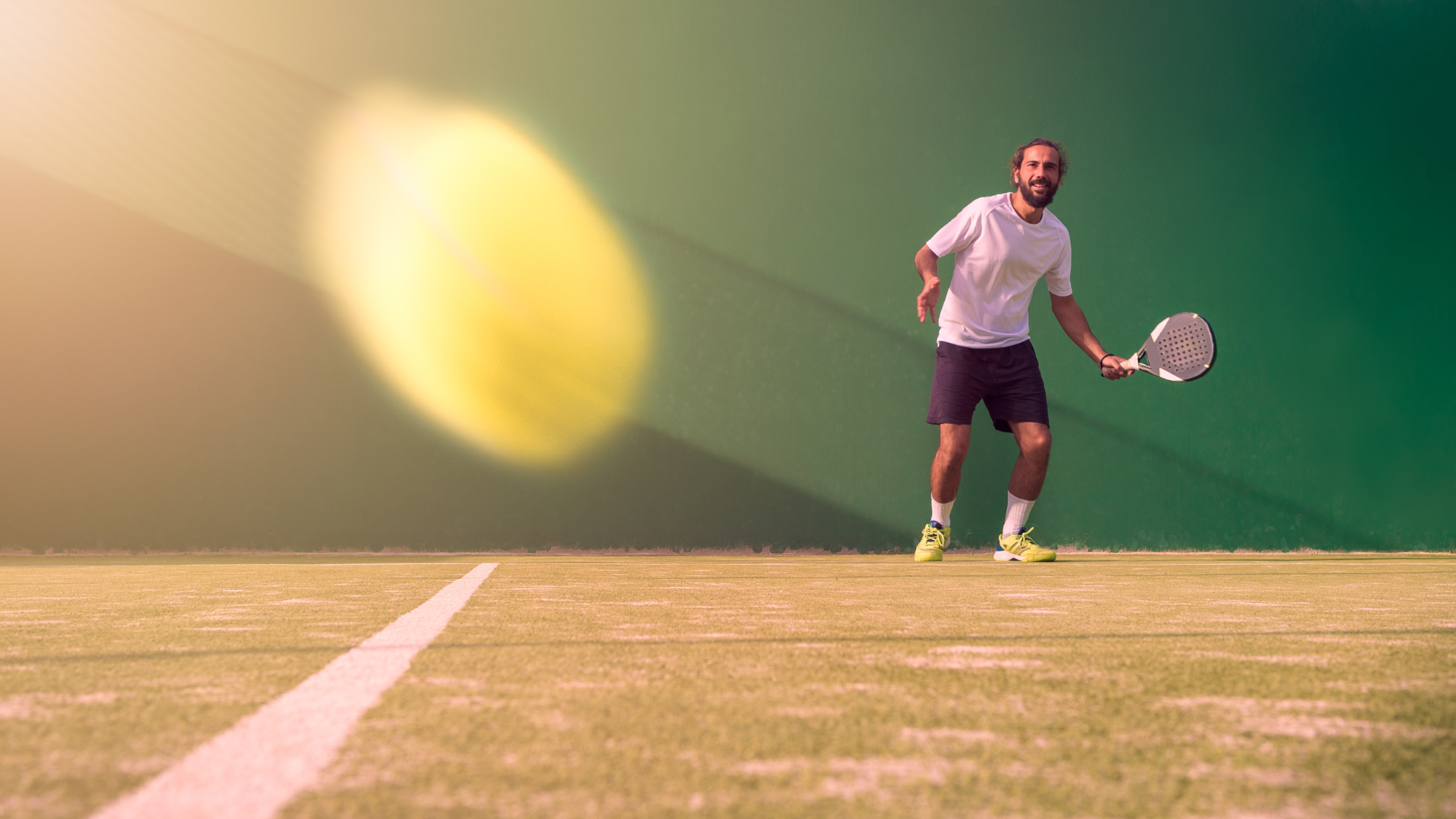 Man playing padel tennis on a grass court with a blurred yellow ball in the foreground.