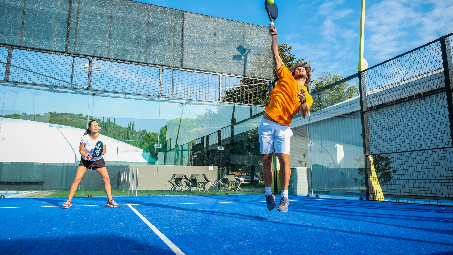 A man and a woman playing padel tennis in the sun on a blue court with glass surround