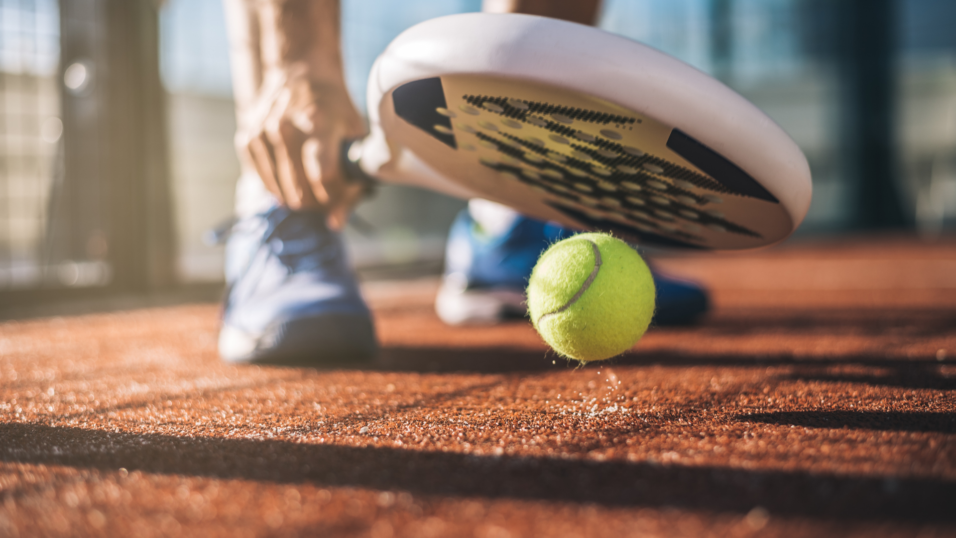 A man bounces a ball on the floor with a padel racket on a terracotta coloured surface